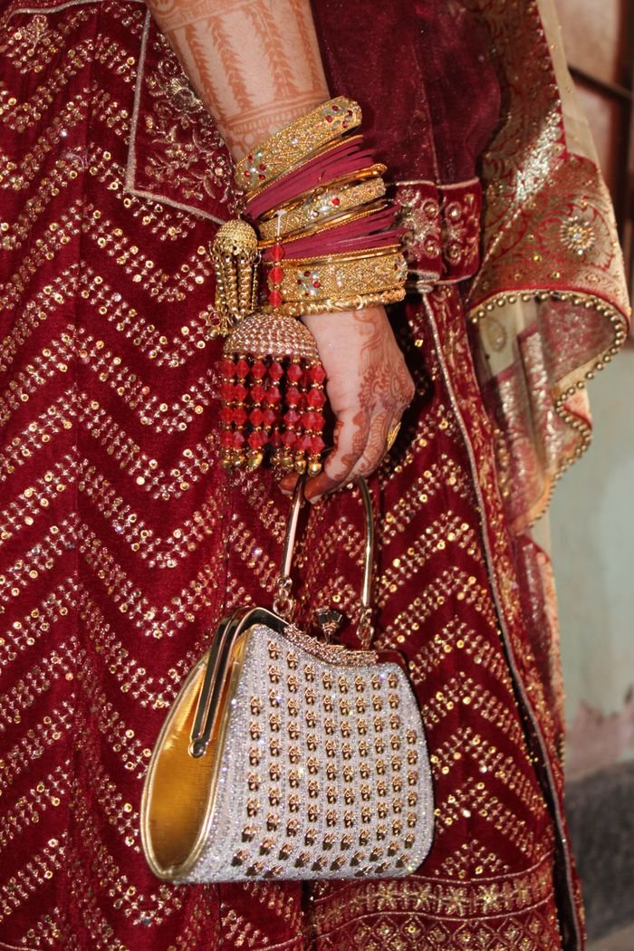 Close-up of a bride's hand in a traditional red saree, showcasing bangles and a decorative purse.