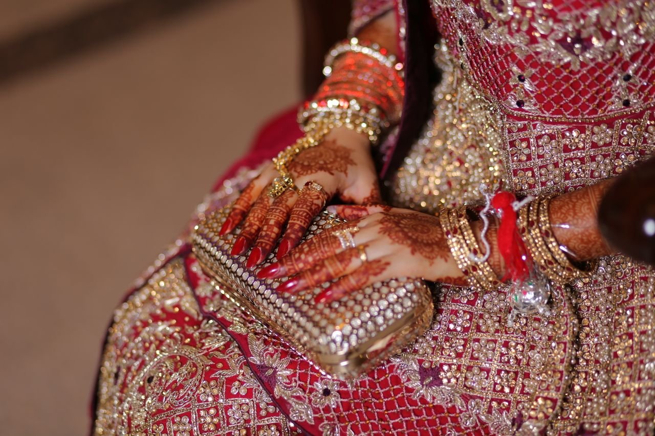 Close-up of henna-decorated bridal hands with jewelry, holding a clutch against a traditional red dress.