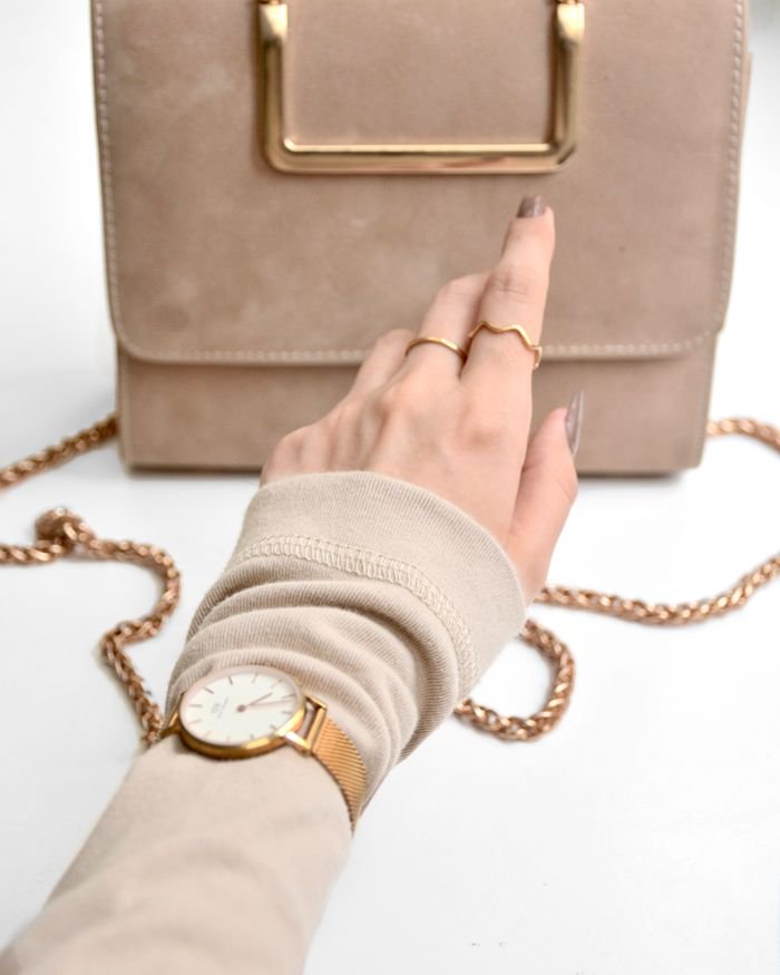 Stylish hand with gold rings, watch, and beige purse against a white background.